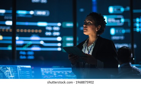 Young Multiethnic Female Government Employee Uses Tablet Computer in System Control Monitoring Center. In the Background Her Coworkers at Their Workspaces with Many Displays Showing Technical Data. - Shutterstock ID 1968875902