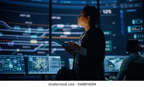 Young Multiethnic Female Government Employee Uses Tablet Computer In System Control Monitoring Center. In The Background Her Coworkers At Their Workspaces With Many Displays Showing Technical Data.