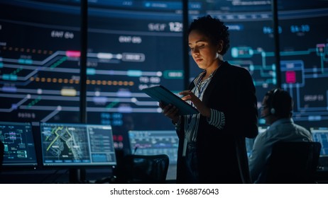 Young Multiethnic Female Government Employee Uses Tablet Computer In System Control Monitoring Center. In The Background Her Coworkers At Their Workspaces With Many Displays Showing Technical Data.