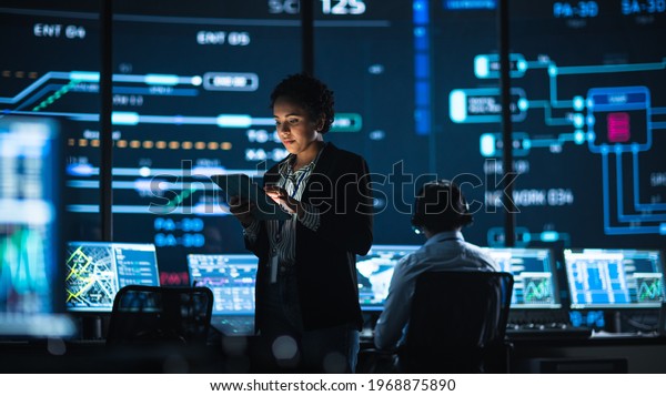 Young Multiethnic Female Government Employee Uses\
Tablet Computer in System Control Monitoring Center. In the\
Background Her Coworkers at Their Workspaces with Many Displays\
Showing Technical Data.