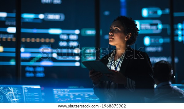 Young Multiethnic Female Government Employee Uses
Tablet Computer in System Control Monitoring Center. In the
Background Her Coworkers at Their Workspaces with Many Displays
Showing Technical Data.