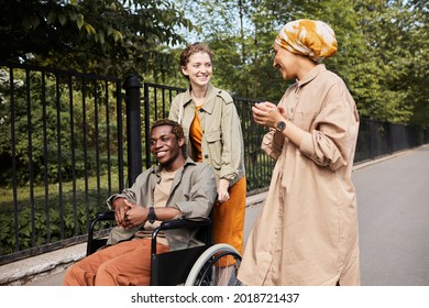 Young multi-ethnic friends talking during stroll: Caucasian woman pushing wheelchair with disabled man while talking to Arabian friend