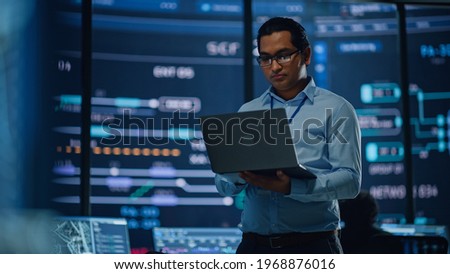 Young Multiethnic Male Government Employee Uses Laptop Computer in System Control Monitoring Center. In the Background His Coworkers at Their Workspaces with Many Displays Showing Technical Data.