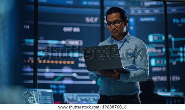 Young Multiethnic Male Government Employee Uses\
Laptop Computer in System Control Monitoring Center. In the\
Background His Coworkers at Their Workspaces with Many Displays\
Showing Technical Data.