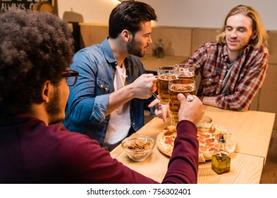 young multiethnic men clinking beer glasses and eating pizza in bar