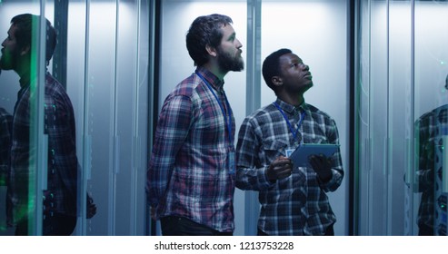 Young multiethnic men using tablet and diagnosing server hardware while walking in data center corridor