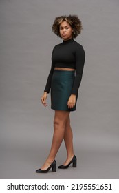 Young Multi-ethnic Woman In Turtleneck Cropped Top And Mini Skirt Posing In Front Of Camera Against Grey Wall During Photo Session