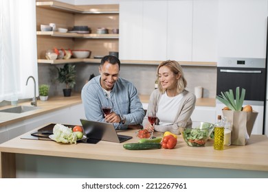 Young Multiracial Couple Communicating With Kids Or Relatives, Having Video Chat On Laptop Computer From Home At Modern Kitchen While Drinking Red Wine.