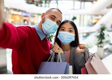 Young multiracial couple in face masks taking selfie together while shopping at huge mall. Millennial diverse family with gift bags wearing protection, keeping safe at mall during covid lockdown - Powered by Shutterstock