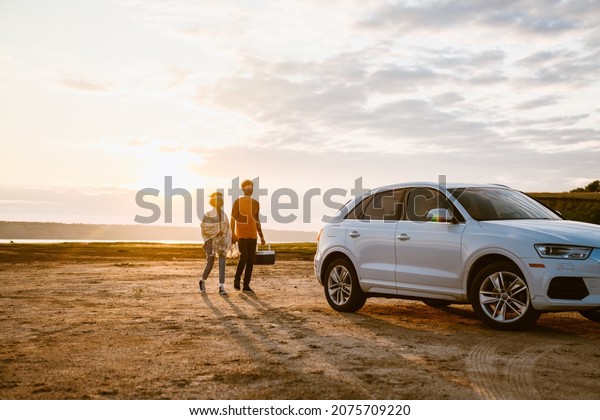Young multiracial couple going with cooler bag\
to their car outdoors