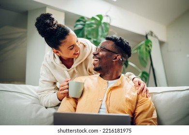 Young Multiracial Couple Relaxing On A Sofa At Home While Using A Laptop. African American Man Holding A Computer And Hispanic Woman Is Hugging Him From Behind With A Cup Of Coffee Or Tea In Her Hand.