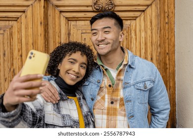 Young multiracial couple smiling and taking a selfie with a smartphone in front of a wooden door, celebrating valentine's day with joy and love - Powered by Shutterstock