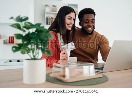 Young multiracial couple smiling, using laptop and credit card for online shopping at home kitchen counter, enjoying e commerce
