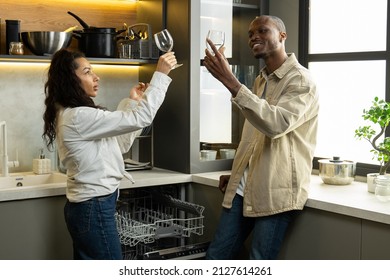 Young Multiracial Couple Takes Clean Wineglasses Out Of Dishwasher In Kitchen. Biracial Woman And African American Man Check Goblets For Stains