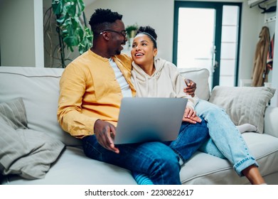 Young multiracial couple using laptop together on sofa at home on the weekend and looking at each other. African american man and hispanic woman browsing internet. Copy space. - Powered by Shutterstock