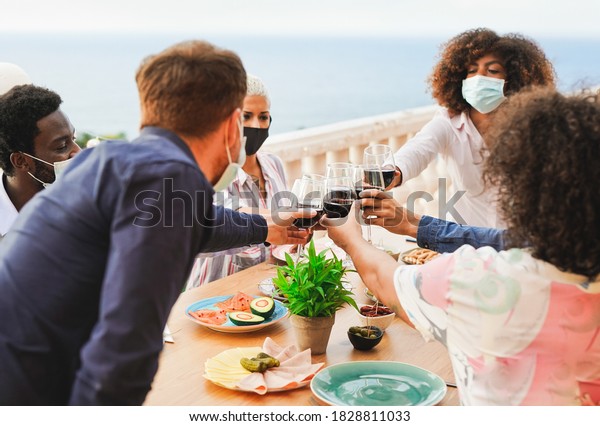 Young\
multiracial people cheering with wine - Dinner outdoor while\
wearing protective face mask for\
coronavirus