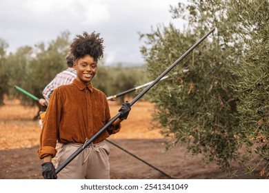 Young, multiracial woman working as olive picker smiles while holding a harvesting tool in an olive grove, her coworker working in the background - Powered by Shutterstock