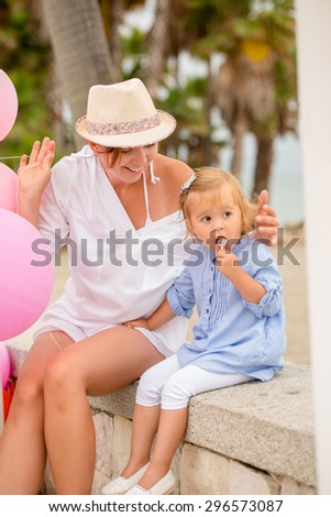 Young Mum in trendy summer clothing playing, with her cute little daughter as they sit together on a wall outdoors at a kids birthday party with pink balloons