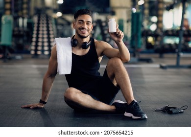 Young Muscular Arab Man Holding Container With Supplement Pills, Middle Eastern Male Athlete Posing At Gym With Blank Pack Of Multivitamins In Hands, Sitting On Floor In Sport Club And Smiling - Shutterstock ID 2075654215