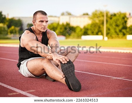 Young muscular athletic runner man stretching and touching his feet on a running court in sitting position before starting of running - Jogging and flexibility concept - Focus on the runner's face