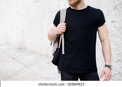 Young Muscular Bearded Man Wearing Black Tshirt And Backpack Posing Outside. Empty White Concrete Wall On The Background. Hotizontal Mockup, Front View.