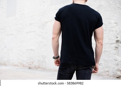 Young muscular bearded man wearing black tshirt and jeans posing in center of modern city. Empty concrete wall on the background. Hotizontal mockup, back view.