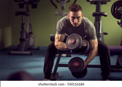 Молодой мускулистый спортсмен тренирует в тренажерном зале, сидя на машине тяжелой атлетики и поднимая два гантеля