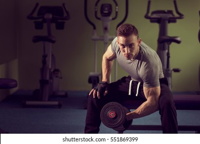 Young Muscular Built Athlete Working Out In A Gym, Sitting On A Weightlifting Machine And Lifting Two Dumbbells