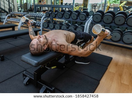 Young muscular fitness man trains pectoral muscles while lying on a bench with dumbbells in his arms at the gym