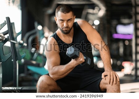 Young Muscular Man Doing Exercises With Dumbbell In Modern Gym, Handsome Athletic Male Making Seated Biceps Curl, Enjoying Workout With Heavy Weights For Muscle Strength, Free Space