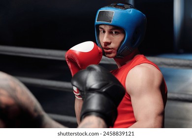 Young muscular man in red vest and boxing gloves and blue face shield looking at his rival before kick during sparring fight on ring - Powered by Shutterstock