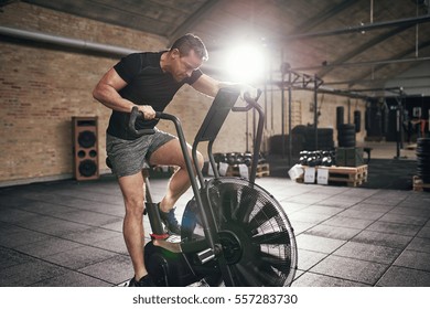 Young Muscular Man In Sportswear Sitting On Cycling Machine And Riding Fast In Modern Gym. 