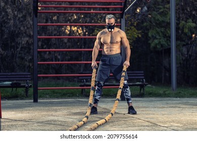 Young Muscular Man Without A Shirt Working Out Outside Using Battle Rope And High Altitude Breathing Simulation Mask, Showing Muscles Concept