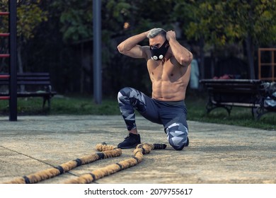 Young Muscular Man Without A Shirt Working Out Outside Using Battle Rope And High Altitude Breathing Simulation Mask, Showing Muscles Concept