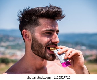 Young Muscular Shirtless Man Eating Protein Bar Outdooron A Balcony In Summer