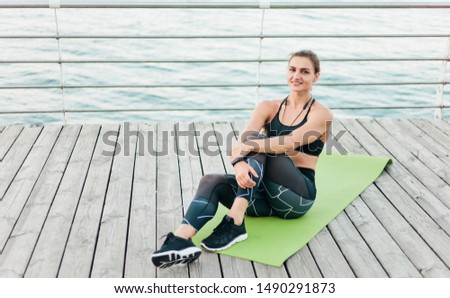 Young muscular woman in sportswear is resting while sitting on mat at beach terrace. Healthy lifestyle concept.