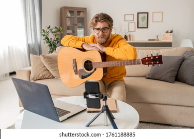 Young Music Teacher Talking To His Audience During Online Lesson Of Playing Guitar While Sitting On Couch In Front Of Smartphone Camera