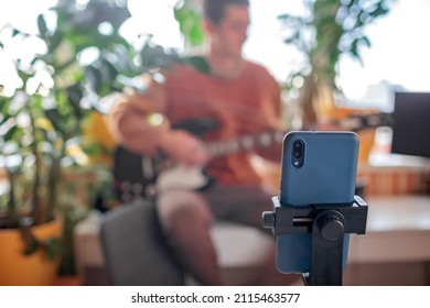 Young musician in cozy sweater sitting on the table and playing electric guitar live in front of the smartphone camera. Online concert during lockdown. Social distance learning. Focus on phone - Powered by Shutterstock