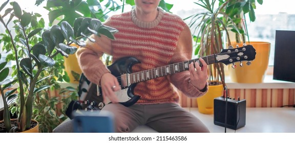 Young musician in cozy sweater sitting on the table and playing electric guitar live in front of the smartphone camera. Online concert during lockdown. Social distance learning. Focus on hand - Powered by Shutterstock