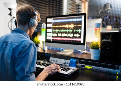 Young musician with headphones pressing keys of piano keyboard while sitting by computer monitor and working with sounds