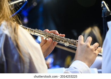 Young Musician Playing Flute On Festival With Band Under On The Stage. Female Musician Playing Flute At Live Classical Concert. Hands Of Musician Playing Instrument With Orchestra. Music Performance.