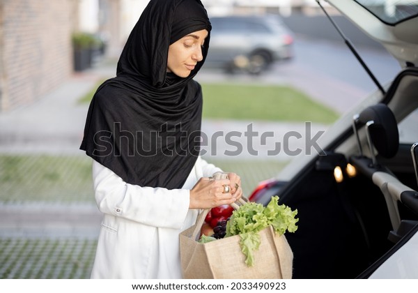 Young muslim\
business woman in hijab going home with groceries, standing with\
shopping bag on the porch of her\
house