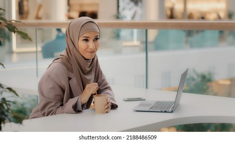 Young Muslim Businesswoman In Hijab Sitting In Office At Desk With Laptop Stirring Coffee In Paper Cup Leisure Happy Arab Girl Reading Pleasant E-mail Thoughtful Shyly Smiled Spends Free Time Thinks