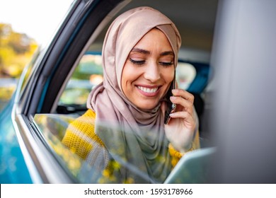 Young Muslim Businesswoman Talking On The Phone In The Back Seat Of The Car And Holding In Hand A Digital Tablet. Beautiful Muslim Business Woman In Car.