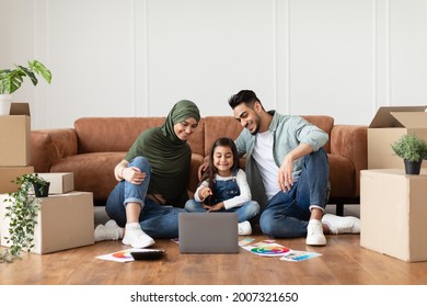 Young Muslim Family Of Three People Using Laptop Sitting On Floor After Moving New Home. Real Estate, House Ownership And Relocation Concept. Parents Browsing Internet, Daughter Pointing At Screen