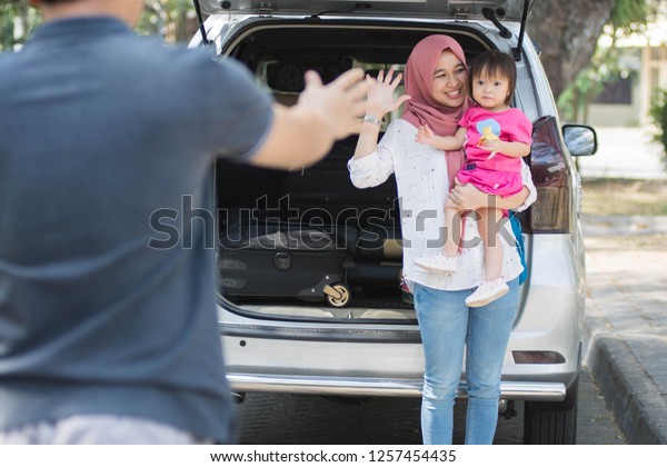 young muslim family, transport, leisure, road\
trip and people concept happy woman and little girl waving at\
father to say goodbye and welcome beside car with trunk open and\
suitcase inside at\
outdoors