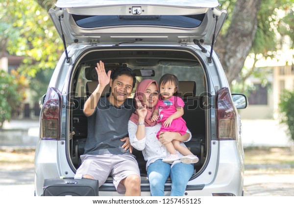 young muslim family , transport,\
leisure, road trip and people concept - happy man, woman and little\
girl sitting on trunk of hatchback car and waving hand\
outdoors