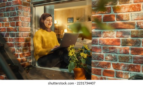 Young Muslim Female in Hijab Using Laptop Computer while Sitting on a Window Sill in Her Stylish Apartment. Creative Manager Working Remotely from Home. Arc Shot from Outside the Window. - Powered by Shutterstock