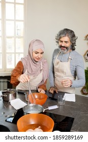 Young Muslim Female Trainee In Hijab Holding Whisker Over Metallic Pan With Water On Electric Stove While Bending Over Table During Cooking