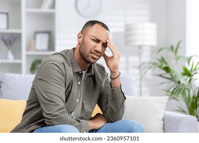 A young Muslim man is suffering from a headache. He sits on the sofa tiredly and massages his hand, grimacing from severe pain and tension. - Powered by Shutterstock